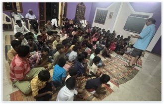 Children seated on woven mats listening to a teacher during an evening children's ministry gathering