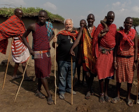 Founder Ray Centanni standing shoulder to shoulder with a group of Maasai men in traditional red shuka cloth in a Kenyan village